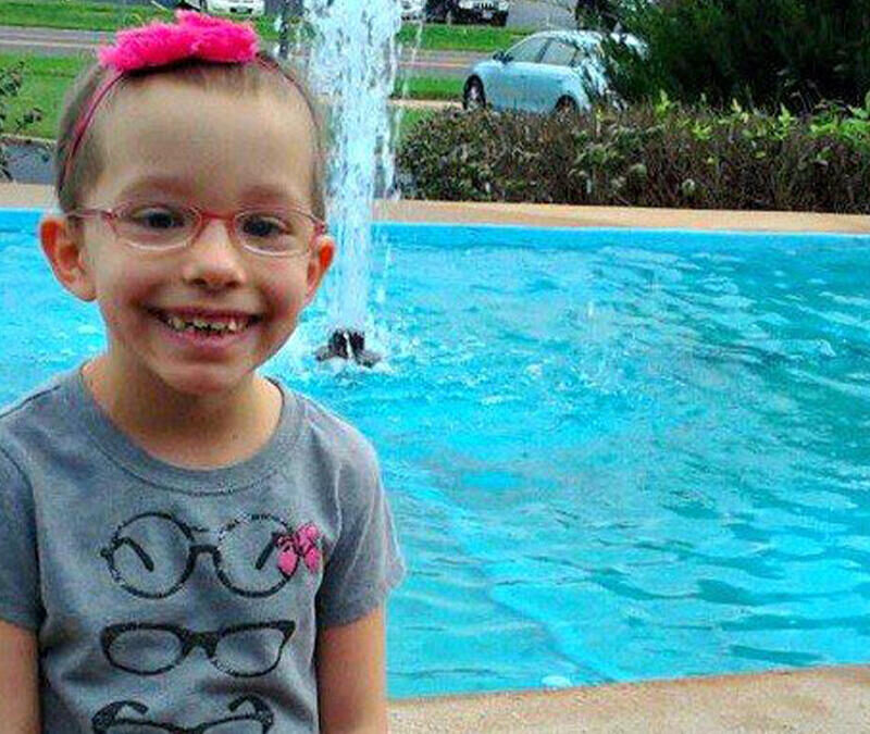 A young girl in glasses smiles in front of a fountain.