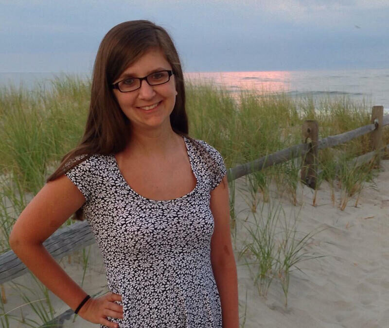 A young woman wearing glasses and a dress standing on the beach.