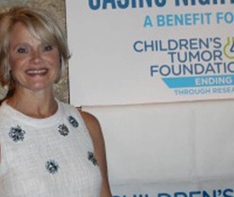 A woman in a white dress standing in front of a children's tumor foundation sign.