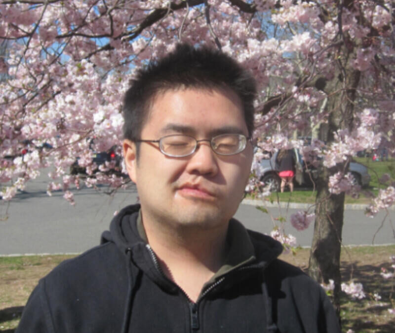 A man with eyes closed standing under a tree with pink flowers.