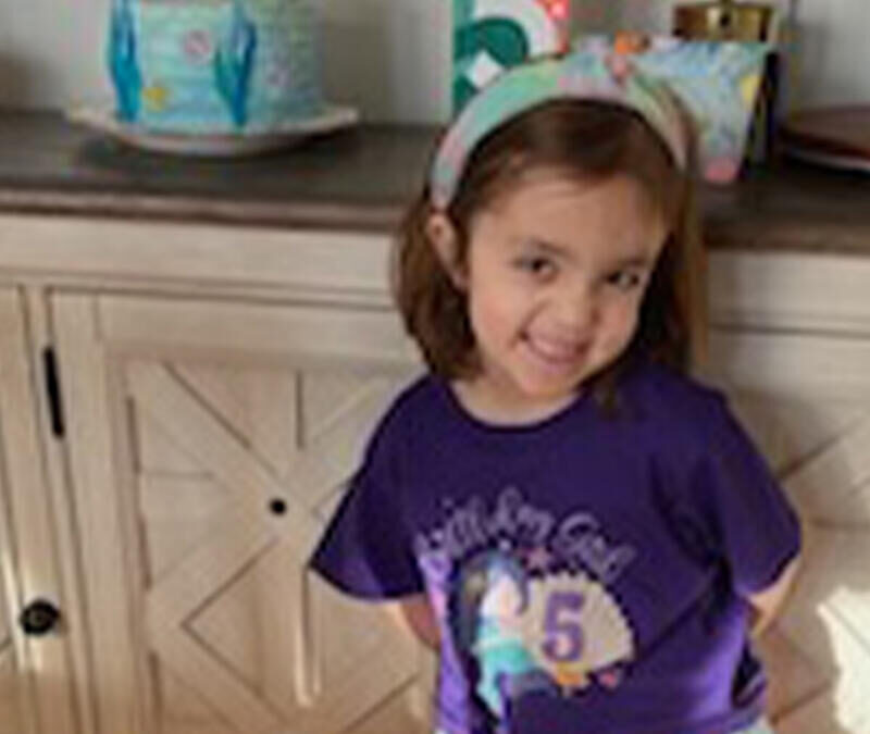 A little girl in a purple shirt standing in front of a cabinet.