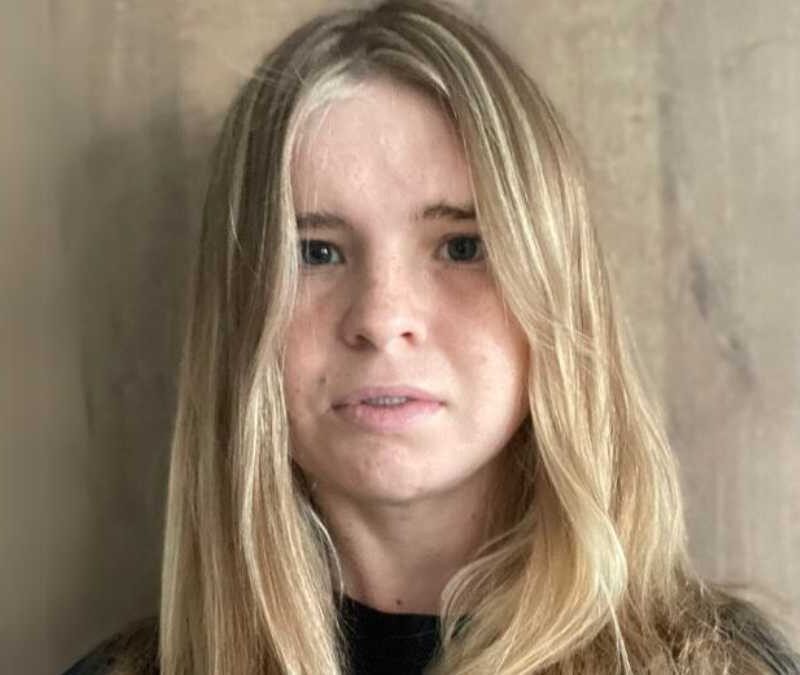 A woman with long blonde hair standing in front of a wooden wall.