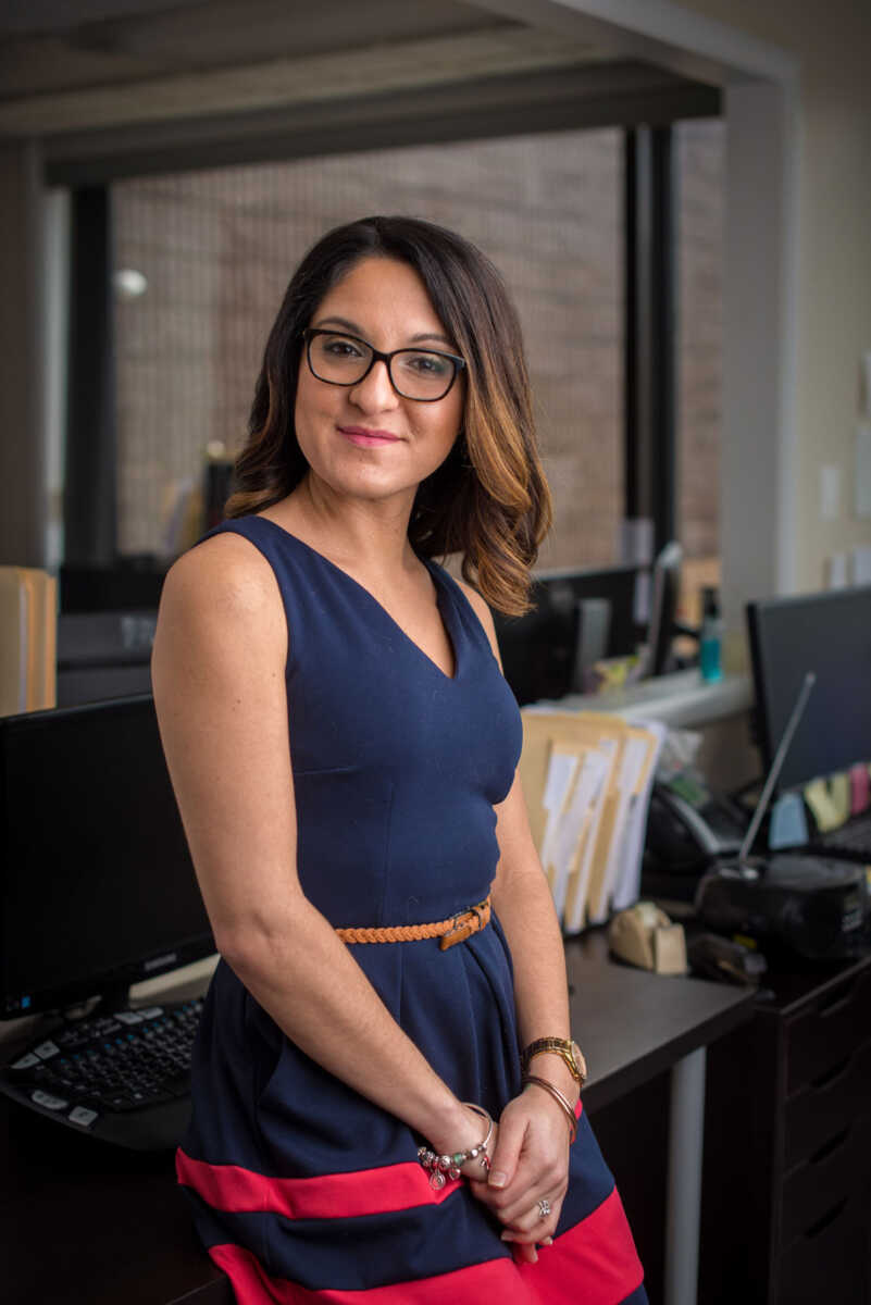 A woman in glasses standing in front of a desk.