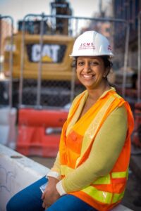 A woman wearing an orange vest and hard hat sits on a construction site.