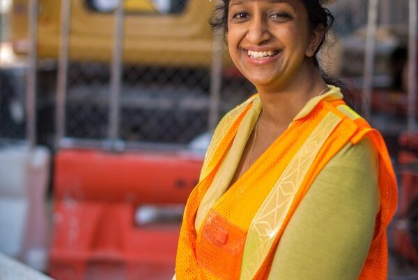 A woman wearing an orange vest and hard hat sits on a construction site.