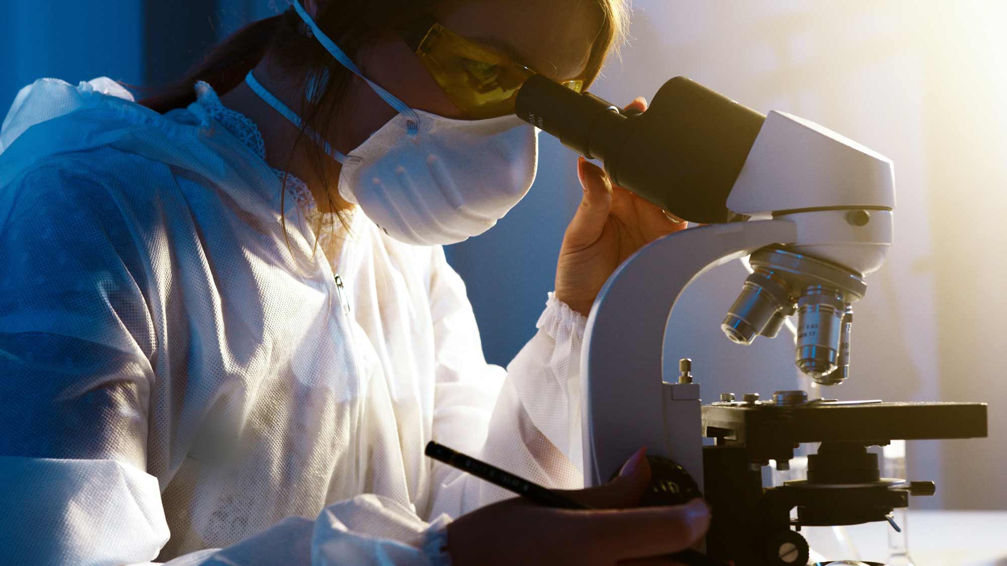 A woman in a white lab coat is looking through a microscope.