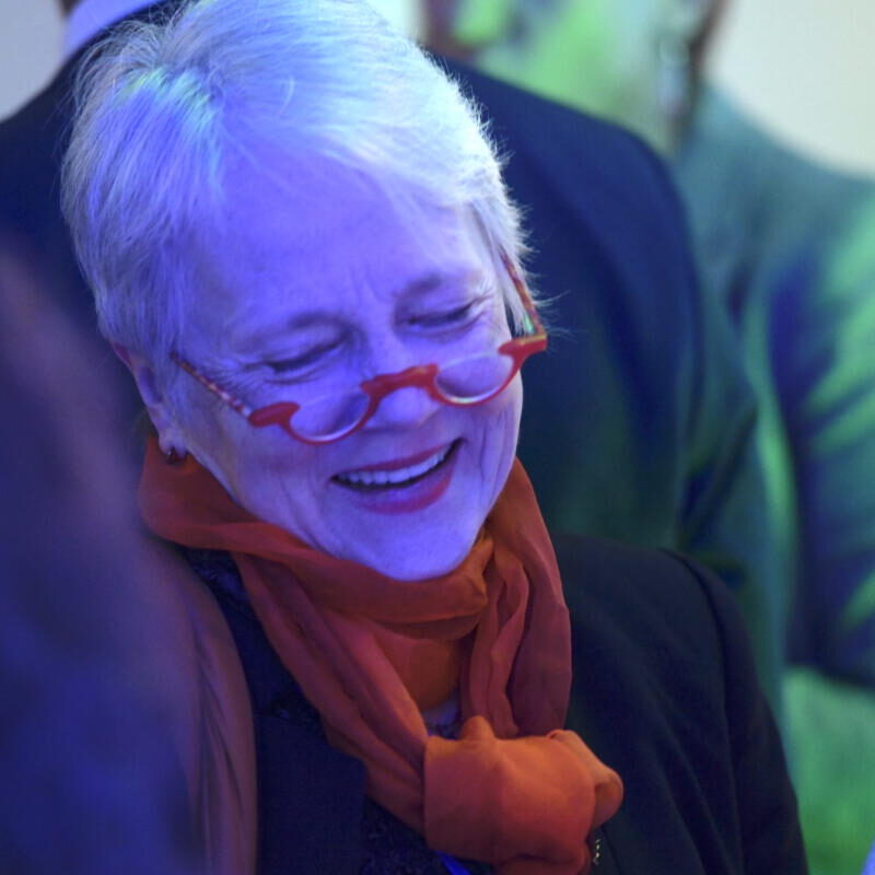 A woman wearing glasses and a scarf smiles at a group of people.