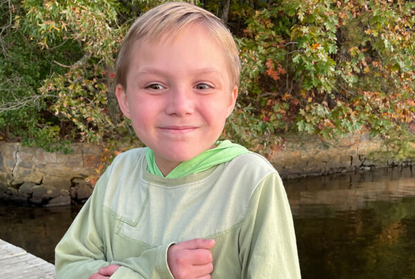 A smiling child in a green hoodie sitting on a dock by the water with autumn foliage in the background.