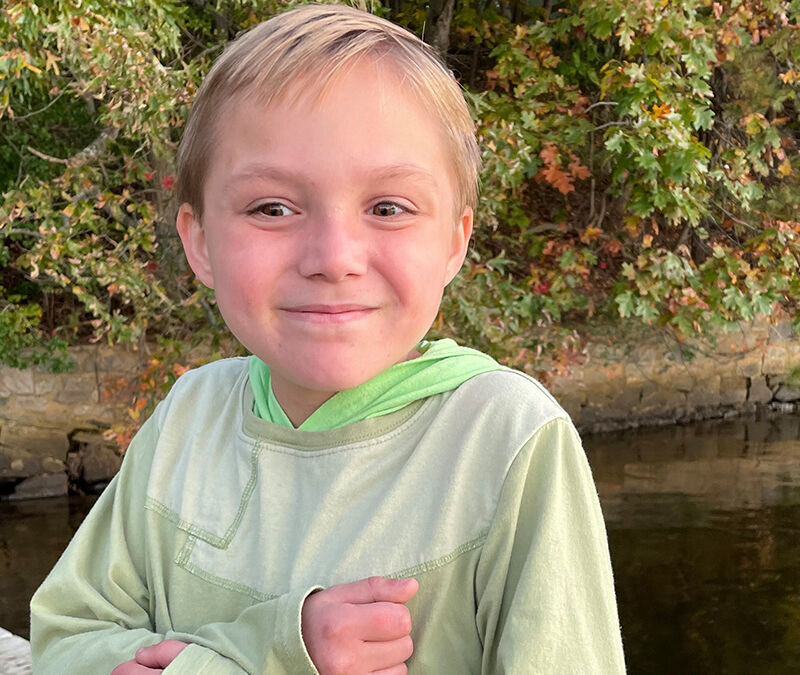 A smiling child in a green hoodie sitting on a dock by the water with autumn foliage in the background.