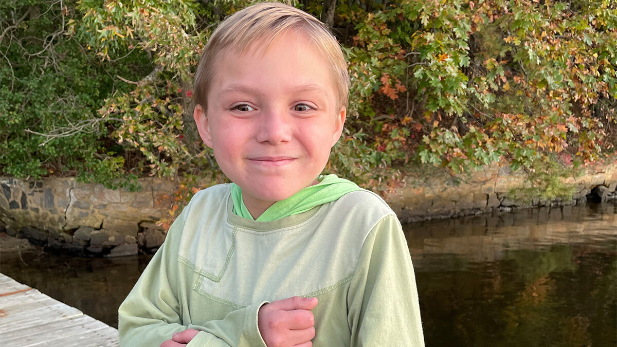 A smiling child in a green hoodie sitting on a dock by the water with autumn foliage in the background.