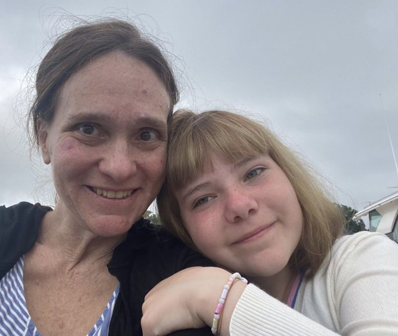 Two people smiling for a selfie with a boat and overcast sky in the background.