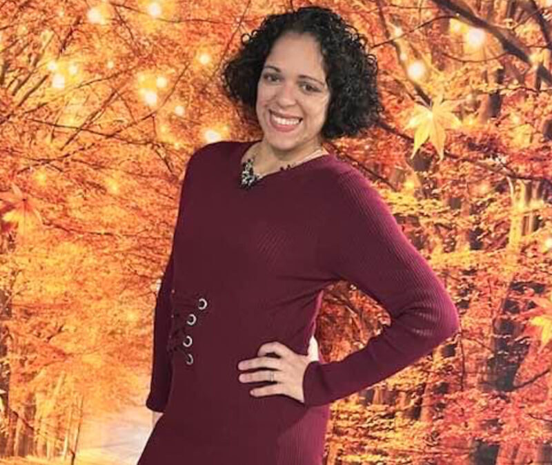 Woman smiling with her hand on her hip, standing in front of a digitally added autumnal forest backdrop.