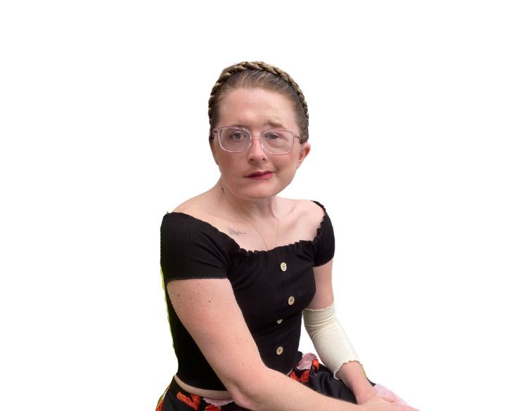 A woman with braided hair and glasses sitting, wearing a black top, floral skirt, and a cast on her left arm, against a white background.