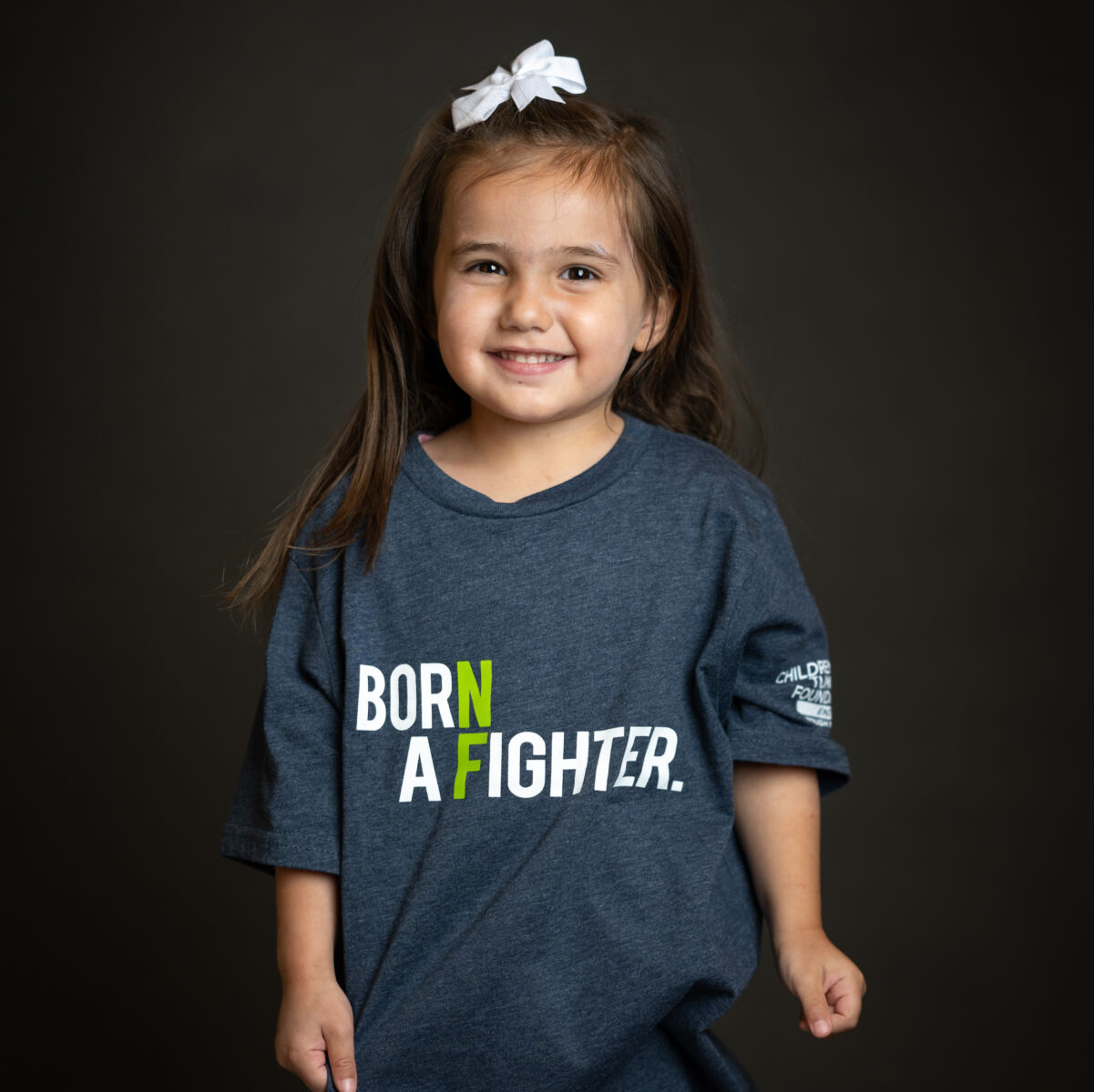 Young girl smiling, wearing a gray t-shirt with "born a fighter." printed on it, standing against a dark background.
