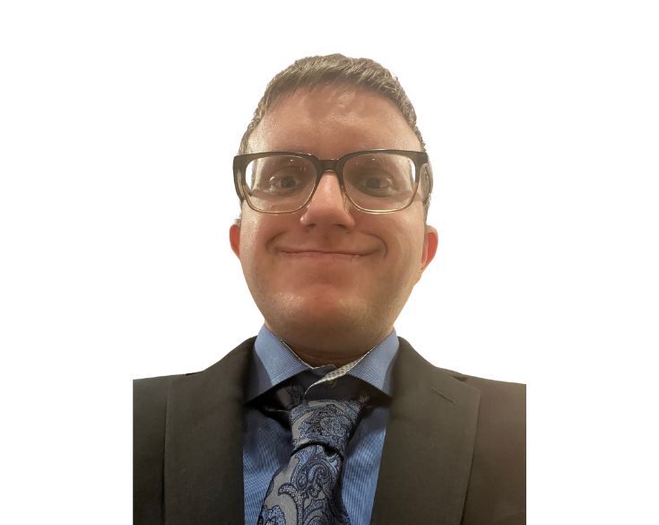 Smiling man with glasses wearing a suit and patterned tie against a white background.