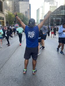 Man celebrating at a marathon, raising arms with a blue "end nf" shirt, spectators and runners in the background.