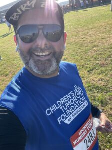 Man in sunglasses and a blue "children's tumor foundation" t-shirt smiles for a selfie at an outdoor event, wearing a race bib numbered 2483.