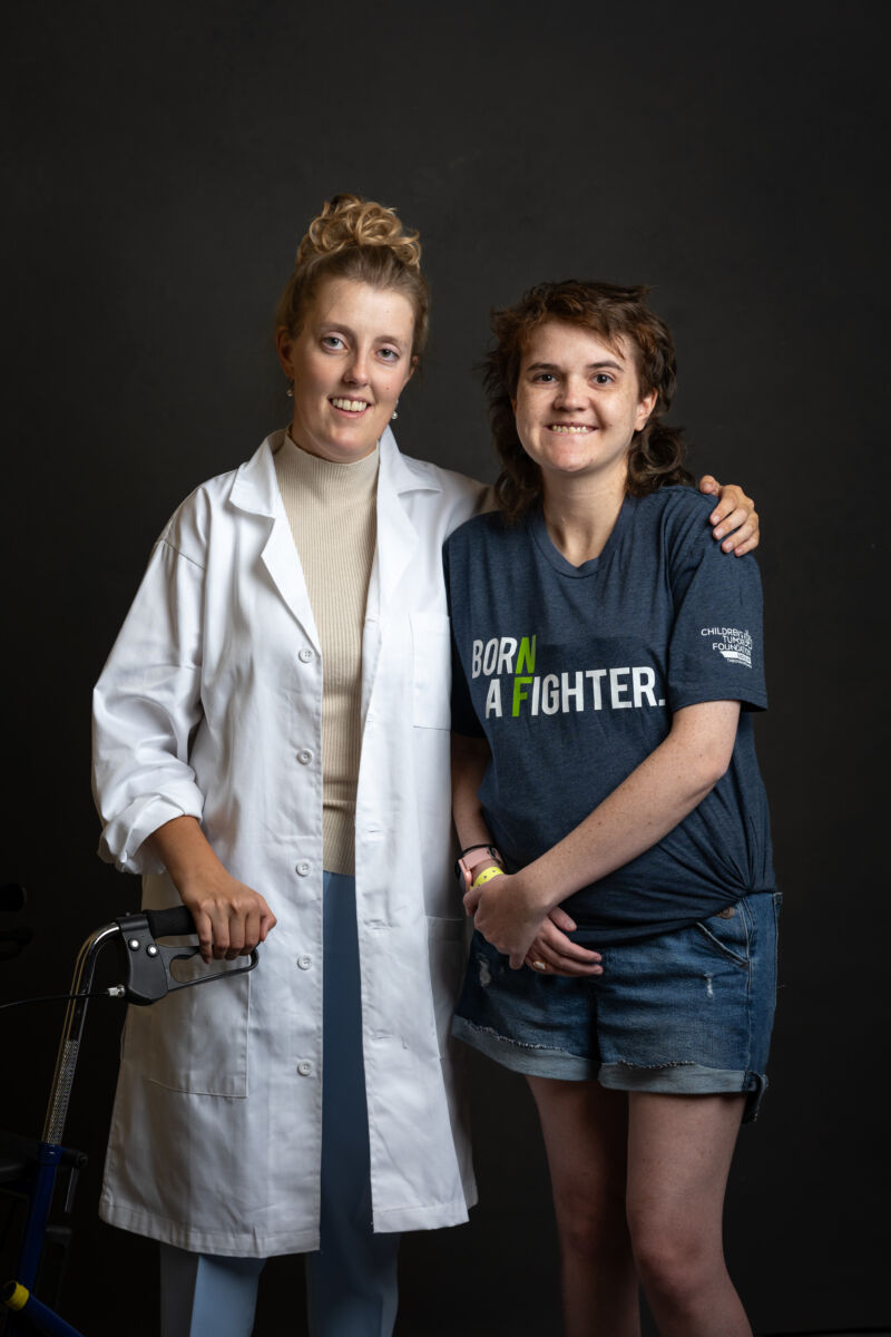 A healthcare professional in a lab coat standing next to a smiling young patient with a walker, both looking at the camera.