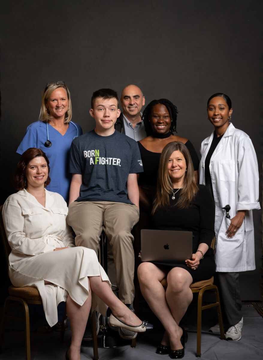 A group of seven individuals, including healthcare professionals and a young man in a "born a fighter" t-shirt, posing for a portrait against a dark backdrop.