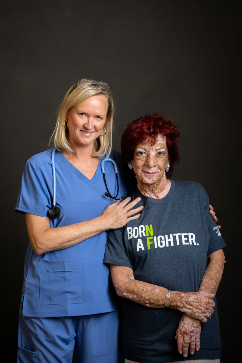 A healthcare worker in blue scrubs stands next to an elderly woman with red hair wearing a 'born a fighter' t-shirt, both smiling for the photo.
