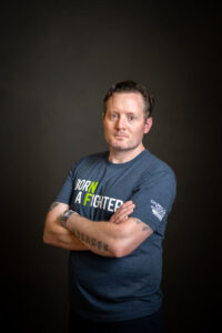 Man with crossed arms wearing a "born a fighter" t-shirt, standing against a dark background, looking directly at the camera.