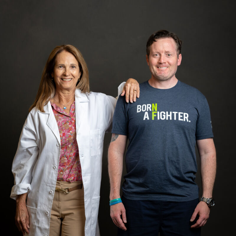 A woman in a lab coat and a man in a "born a fighter" t-shirt smiling, standing side by side against a dark background.