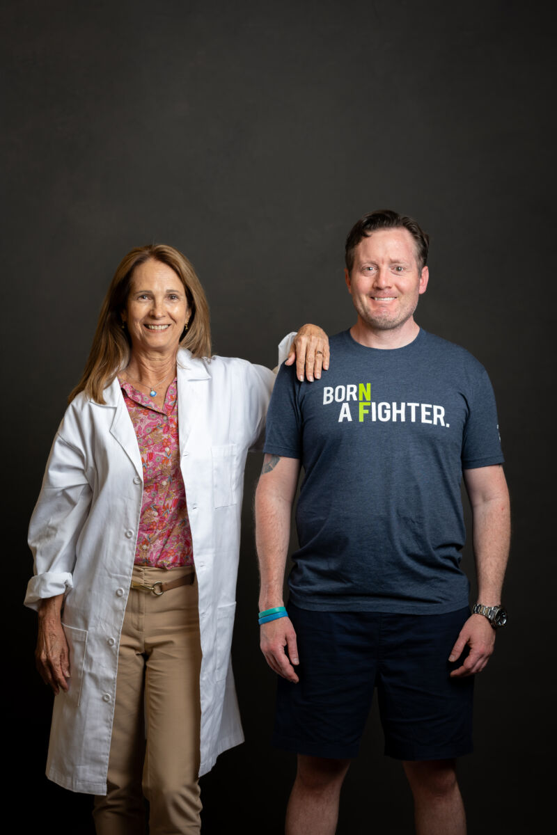 A woman in a lab coat and a man in a "born a fighter" t-shirt smiling, standing side by side against a dark background.