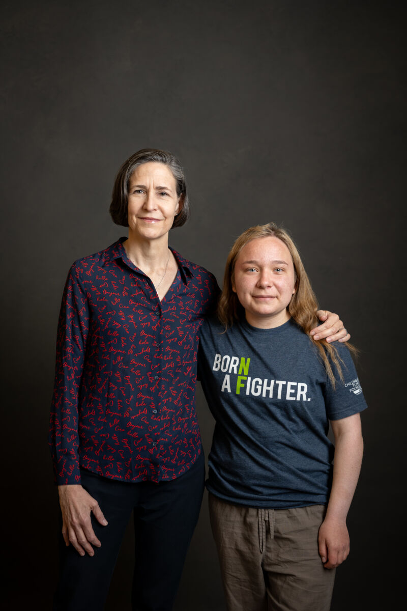 Two individuals standing side by side against a dark backdrop, the taller person wearing a patterned shirt and the shorter person in a t-shirt with the inscription "born a fighter.