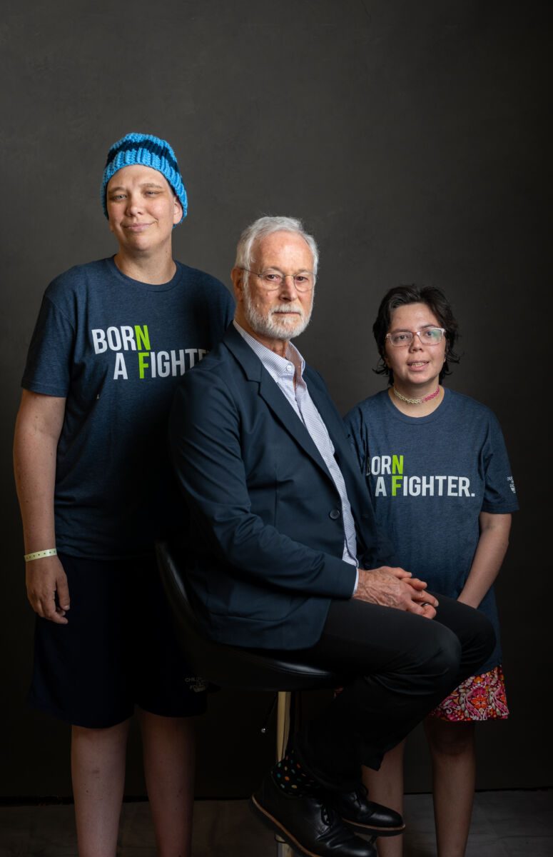 An older man seated on a stool, flanked by two younger individuals standing, all posing for a portrait against a dark background.