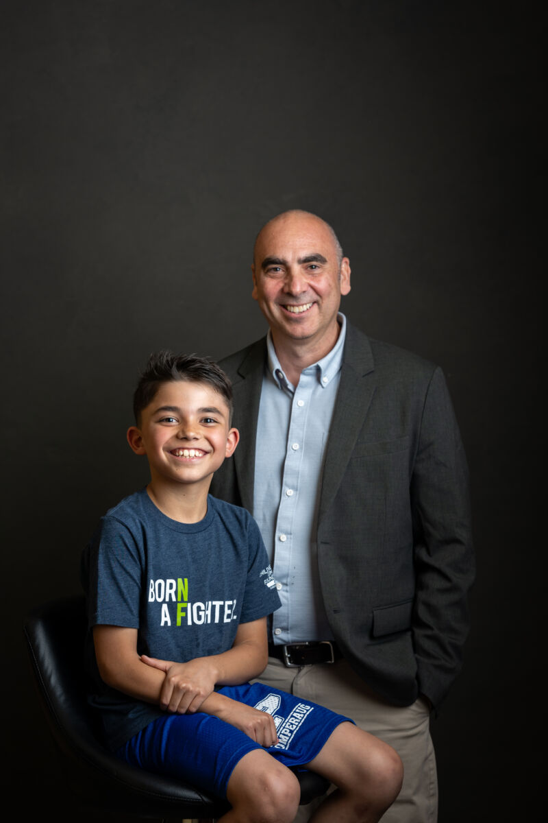 A smiling boy sitting on a chair with a man standing beside him against a dark background.