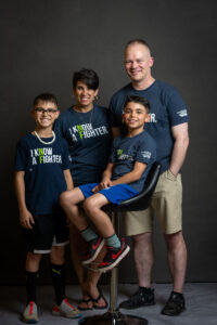A family of four, smiling in matching t-shirts, posing together with one child and one adult seated on a stool.