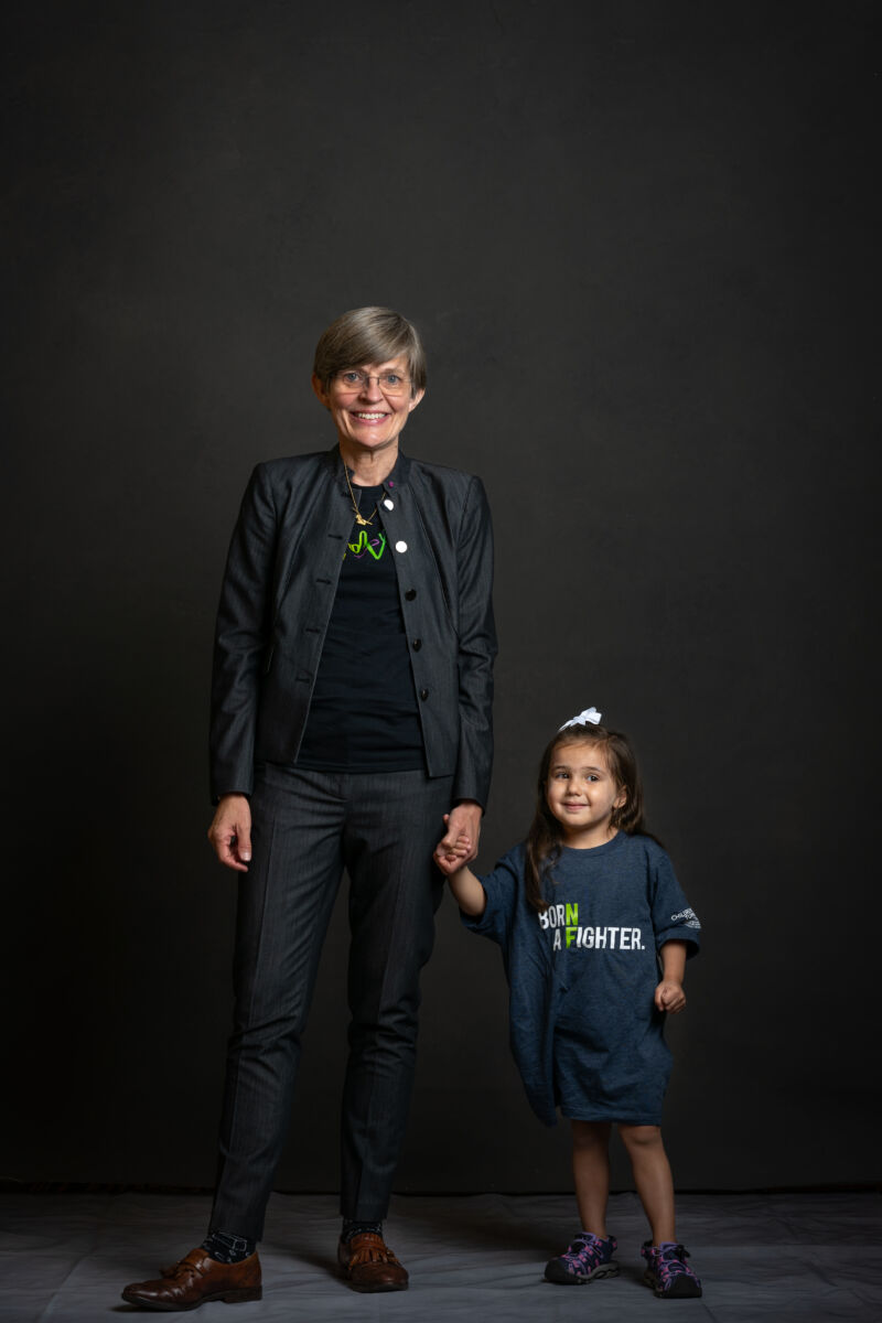A woman in a suit stands holding hands with a small child wearing a "sunburn fighter" t-shirt, both smiling against a dark backdrop.