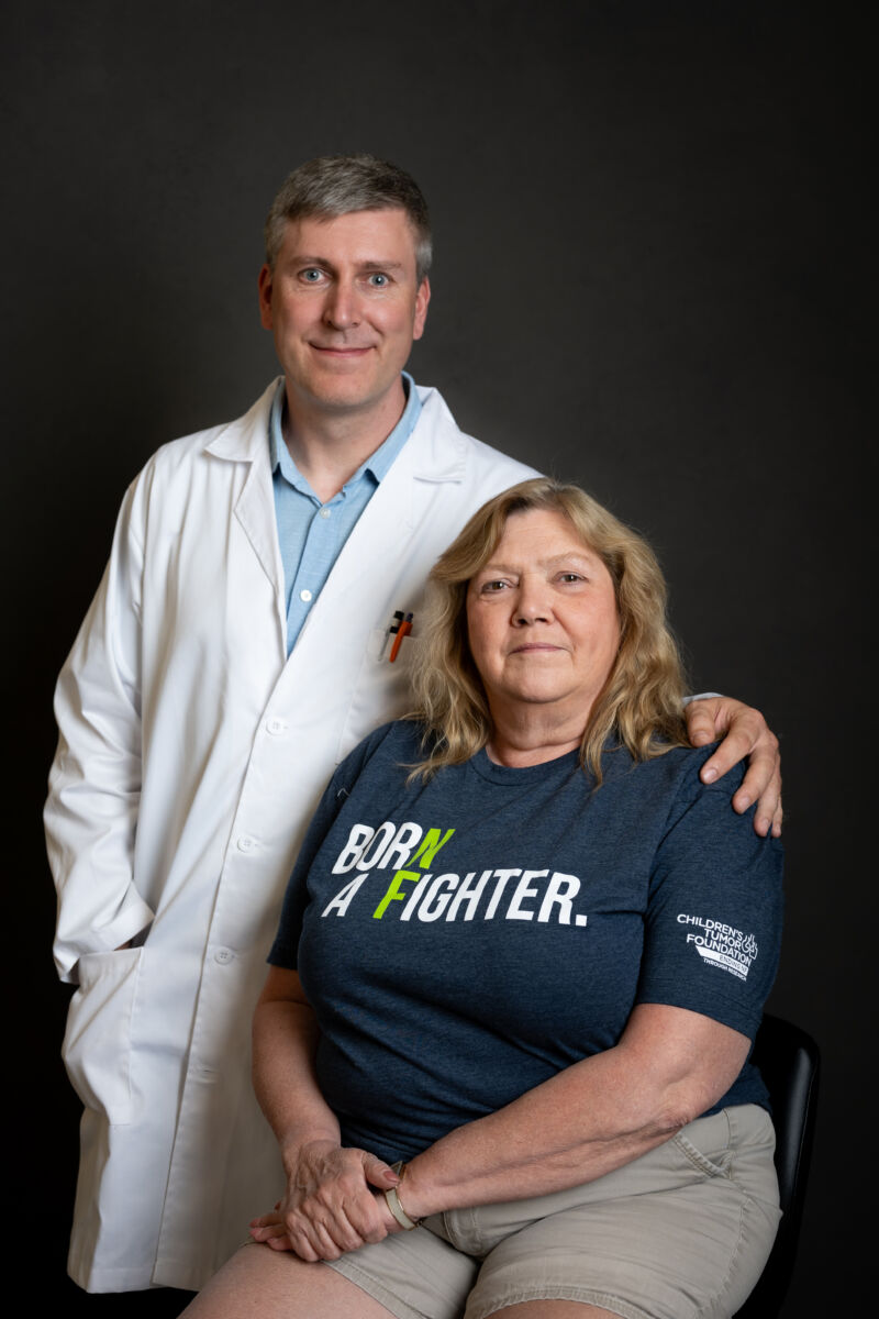 Doctor standing behind seated female patient, both smiling at the camera against a black background.