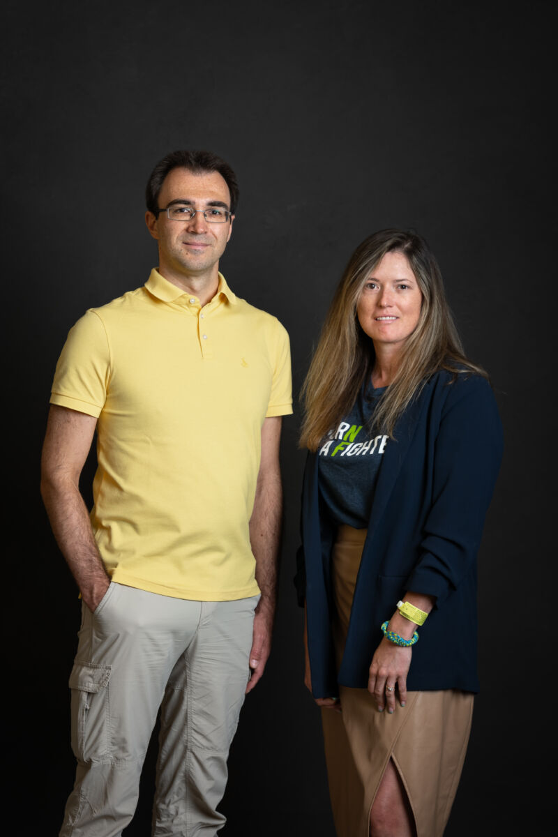 Two individuals posing for a portrait against a black backdrop; the person on the left is wearing a yellow polo shirt and khaki pants, and the person on the right is dressed in a dark blazer over a graphic t-shirt with light brown pants.