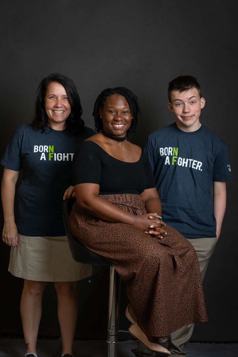 Three individuals smiling at the camera, two wearing t-shirts with the text "born a fighter" against a black background.