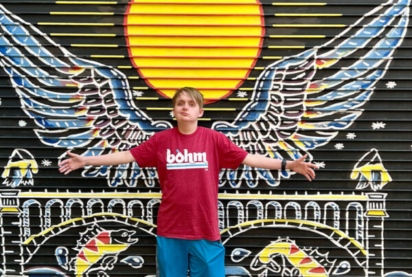 A teenage boy stands with arms outstretched in front of a colorful mural of wings and a sun, wearing a casual t-shirt and shorts.