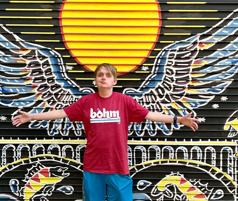 A teenage boy stands with arms outstretched in front of a colorful mural of wings and a sun, wearing a casual t-shirt and shorts.