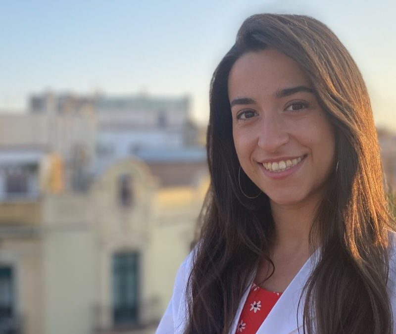 A smiling woman with long hair outdoors, with buildings in the background bathed in warm sunlight.