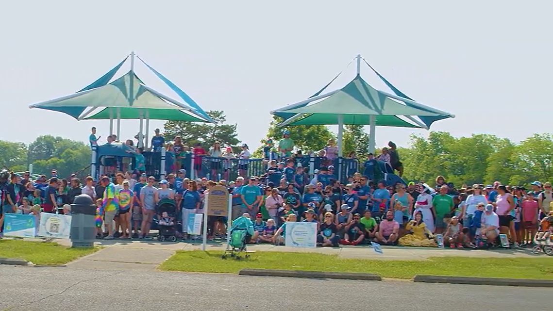 A crowd of spectators gathered under green and white pavilions at an outdoor event on a sunny day.