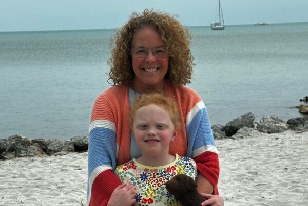 A woman and a young child smiling on a beach with a sailboat in the distance. the child holds a teddy bear.