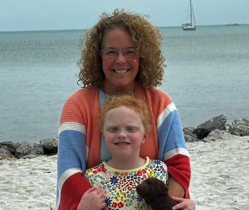 A woman and a young child smiling on a beach with a sailboat in the distance. the child holds a teddy bear.