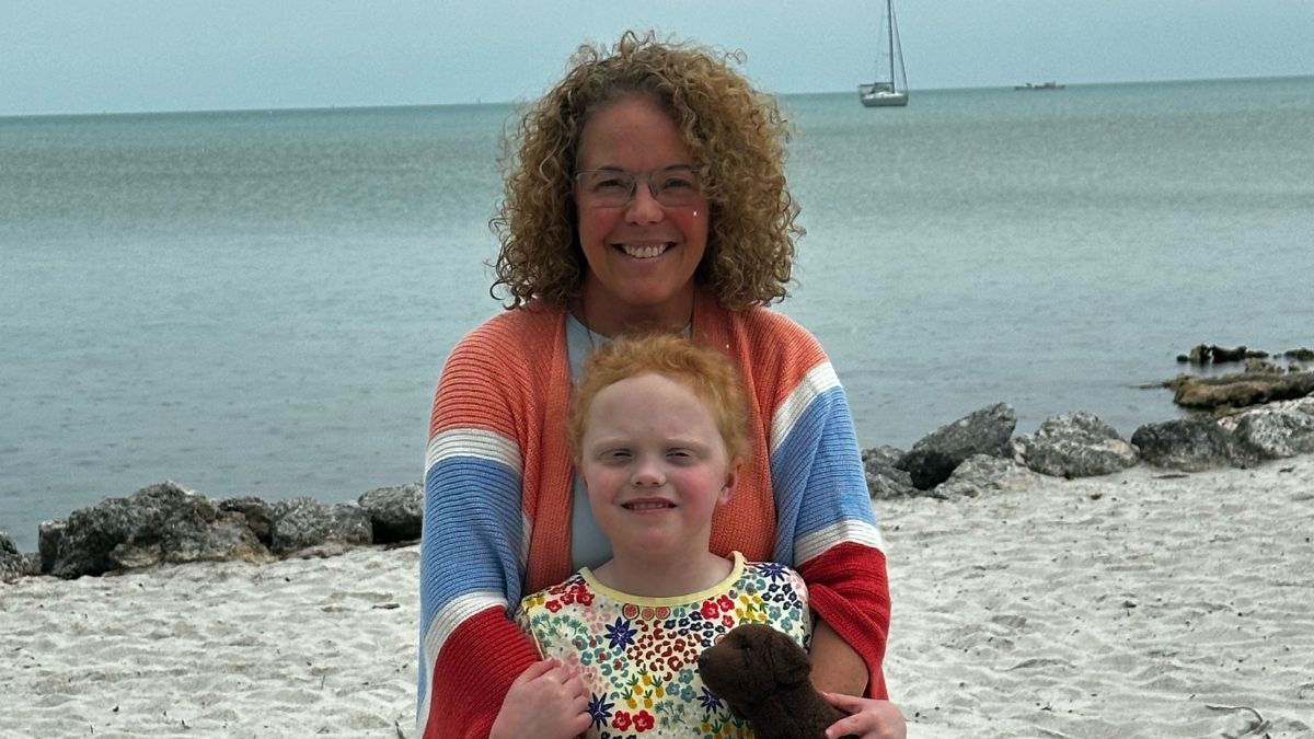 A woman and a young child smiling on a beach with a sailboat in the distance. the child holds a teddy bear.
