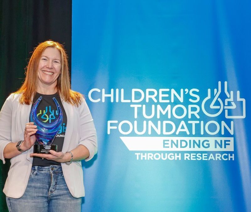 Woman smiling and holding an award at the children's tumor foundation event, with a banner reading "ending nf through research" in the background.