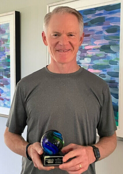 A smiling man holding a trophy with a colorful glass top, standing in a room with abstract art in the background.