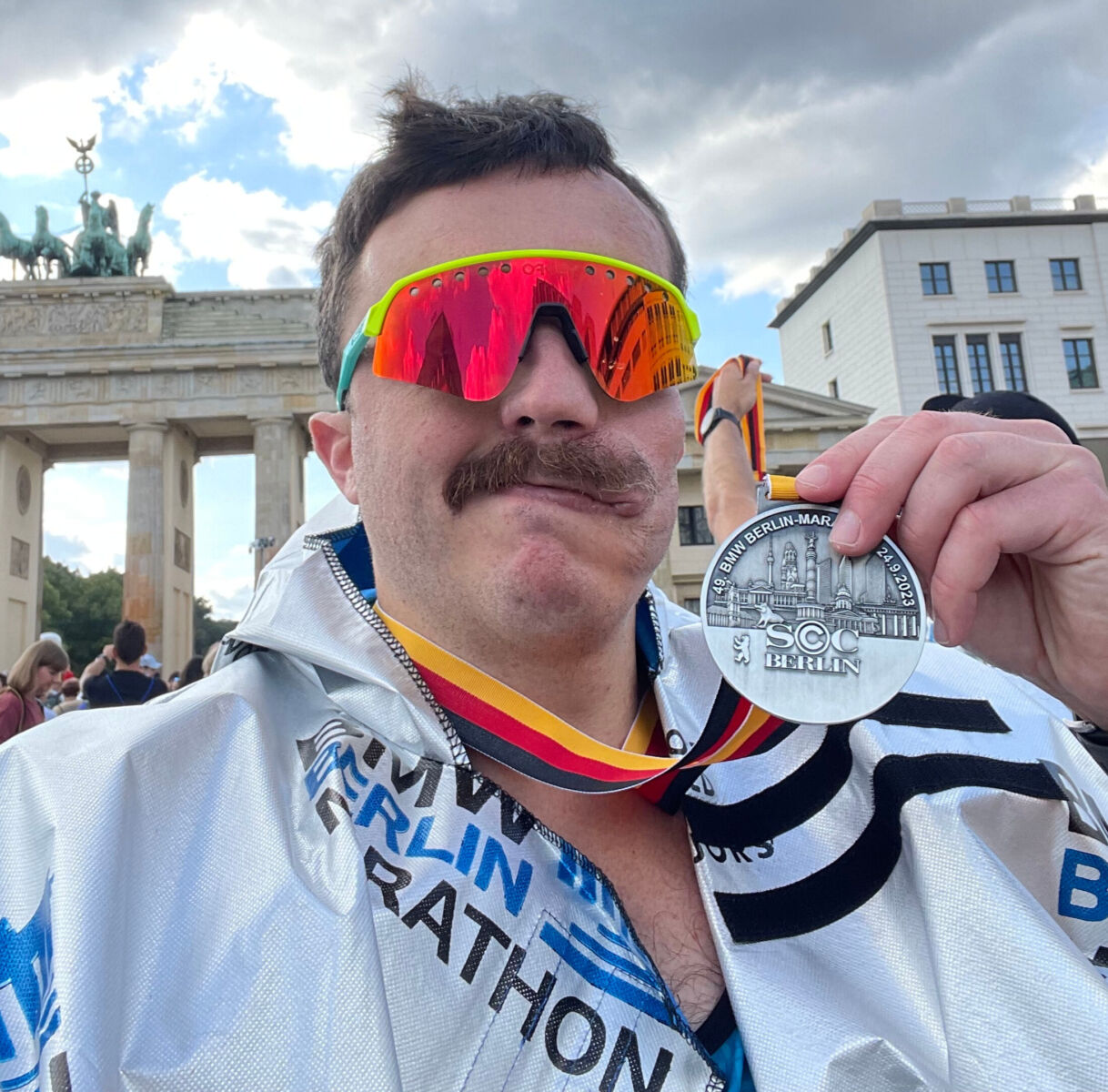 A man with a mustache wearing colorful sunglasses and a marathon medal standing in front of the brandenburg gate.