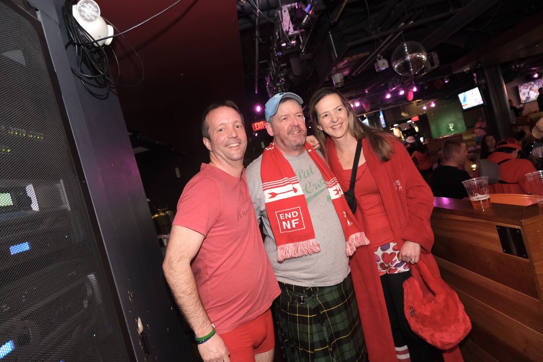 Three adults smiling at a bar event, one in a red t-shirt and shorts, another in a cap and plaid kilt, and the third in a red jacket, holding drinks.