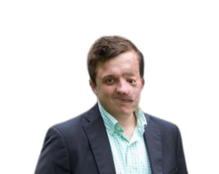 A man with a mustache wearing a dark blazer over a checked shirt, standing against a blurred white background.