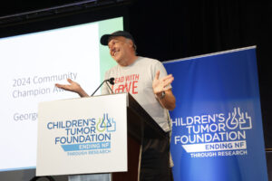 A man speaking at a podium with a "children's tumor foundation" banner, gesturing with his hands, during a presentation for the 2024 community champion award.