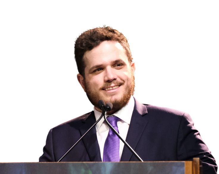A man with a beard, wearing a suit and tie, smiling while standing at a podium.