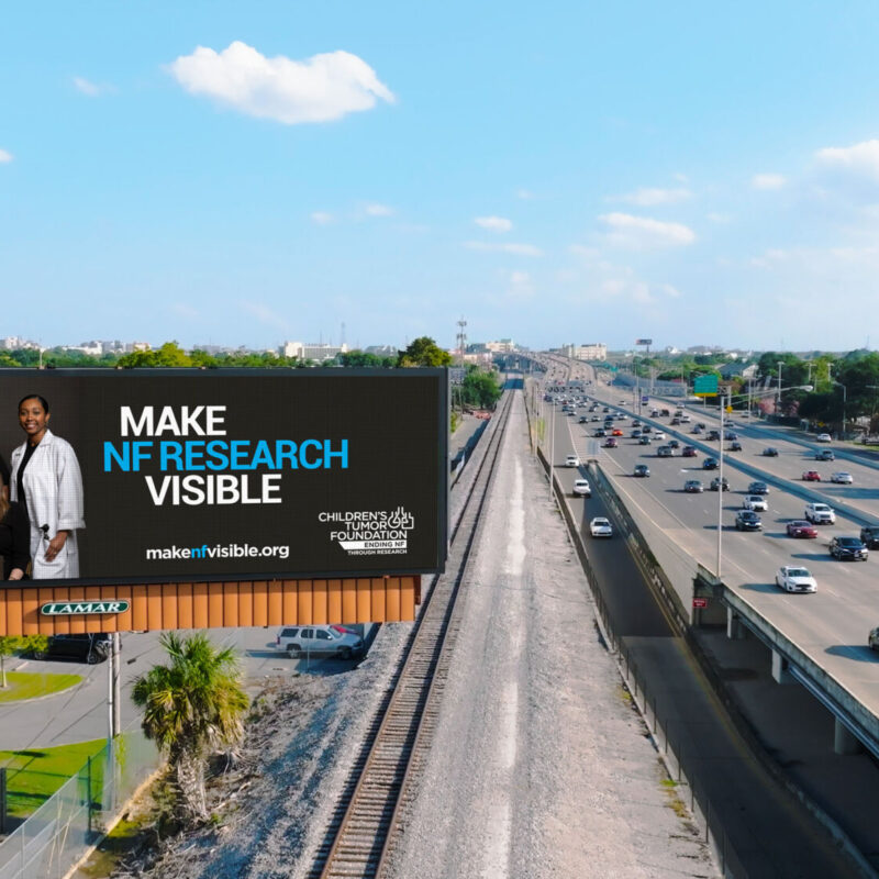 Billboard featuring a group of diverse people with the text "make nf research visible" beside a busy highway with city skyline in distance.